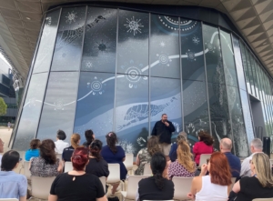 An Aboriginal man in black shirt speaks into a microphone to an audience sat in white chairs in front of a giant mural on the windows of a building